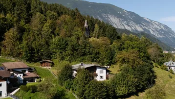 Idyllisches Haus in Hanglage, umgeben von Wald und Bergen unter blauem Himmel.