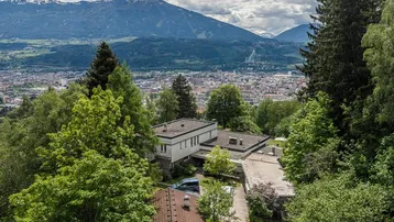 Luftaufnahme eines modernen Hauses mit Flachdach, umgeben von Bäumen, mit Stadt- und Bergblick.