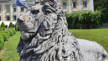 Große steinerne Löwenstatue im Vordergrund eines prächtigen historischen Gebäudes mit gepflegtem Garten.