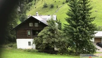 Traditionelles Holzhaus in alpiner Landschaft mit grünen Wiesen und hohen Bäumen.