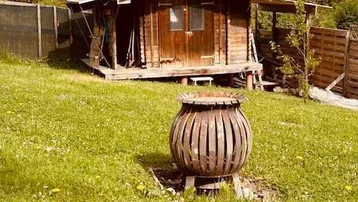 Kleines Holzhaus im Grünen mit Feuerstelle und umgebendem Gartenbereich unter blauem Himmel.
