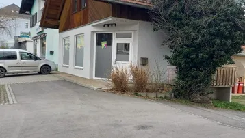 Gebäudeansicht mit Satteldach, weißer Fassade im Erdgeschoss und Holzverkleidung im Obergeschoss, umgeben von einer gepflasterten Fläche.
