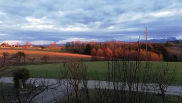 Weitläufiger Landschaftsblick mit grünen Feldern, Bäumen und Bergen im Hintergrund bei Sonnenuntergang.