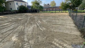 Großes, geräumtes Baugrundstück mit vorbereitetem Untergrund und einem Haus im Hintergrund.