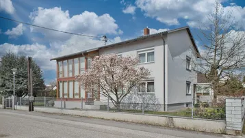 Freistehendes Einfamilienhaus mit heller Fassade, gepflegtem Vorgarten und blühendem Baum an der Straße.