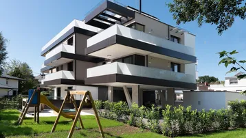 Modernes Mehrfamilienhaus mit gestuften Balkonen, Spielplatz und gepflegter Grünfläche unter blauem Himmel.