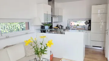 Moderne, halboffene Küche mit weißen Schränken, Holzboden und viel Tageslicht.