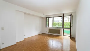 Helles Zimmer mit Parkettboden, weißen Wänden und Zugang zu einem Balkon mit grüner Aussicht.