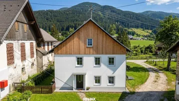 Traditionelles Haus mit weißer Fassade und Holzverkleidung, umgeben von grüner Landschaft und Bergen.