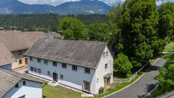 Luftaufnahme eines Wohnhauses mit Satteldach und umliegender grüner Landschaft sowie Bergen im Hintergrund.