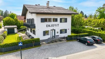 Weißes Haus mit dunklem Dach, Fensterläden und gepflastertem Vorplatz mit Parkplätzen.