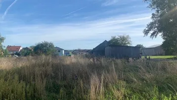 Weitläufiges Grundstück mit hohem Gras und ländlicher Bebauung im Hintergrund unter blauem Himmel.