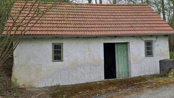 Altes, einfaches Gebäude mit verputzten Wänden, einem Ziegeldach und grünen Holztüren, umgeben von Bäumen.
