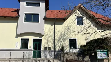 Gelbe Fassade eines Gebäudes mit grauem Anbau und rotem Ziegeldach unter blauem Himmel.