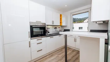 Moderne Küche mit weißen Hochglanzfronten, integriertem Backofen und Blick auf die Berge.