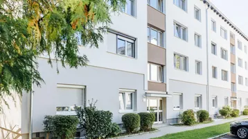 Mehrstöckiges Mehrfamilienhaus mit weißer und grauer Fassade, umgeben von gepflegten Grünflächen.