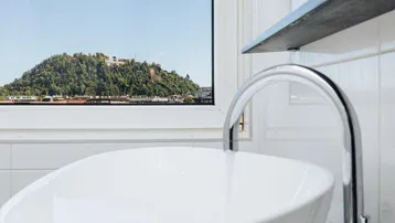 Modernes Badezimmer mit Aufsatzwaschbecken, weißen Fliesen und Blick auf einen bewaldeten Hügel.