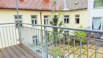 Heller Balkon mit Holzboden und Blick auf den begrünten Innenhof, ideal für sonnige Stunden.