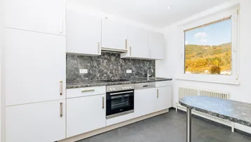 Moderne Küche mit weißen Schränken, dunkler Arbeitsplatte und einem Fenster mit Blick auf die herbstliche Landschaft.