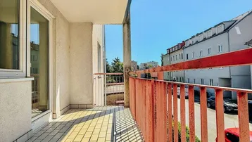 Heller Balkon mit Fliesenboden und rotem Geländer, bietet einen sonnigen Blick auf die umliegende Straße.