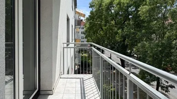 Heller Balkon mit Fliesenboden, Rollläden und Blick auf die grüne Baumreihe.