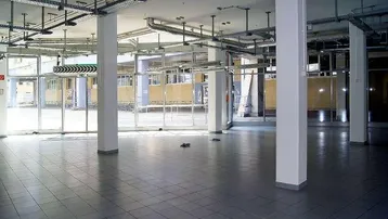 Geräumiger, leerstehender Gewerberaum mit großen Fenstern, Fliesenboden und sichtbaren Lüftungsrohren an der Decke.