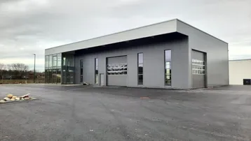 Modernes Gewerbegebäude mit grauer Fassade, großen Fenstern und einem weitläufigen Parkplatz unter bewölktem Himmel.