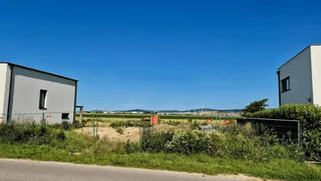 Moderne Gebäude flankieren ein unbebautes Grundstück mit Blick auf Felder und Windräder.