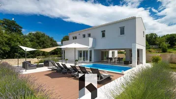 Moderne Villa mit Pool, Holzterrasse und Liegestühlen unter blauem Himmel.
