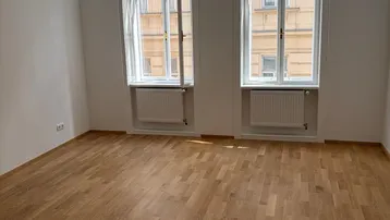 Helles Zimmer mit zwei großen Fenstern, Holzboden und Blick auf die gegenüberliegende Fassade.