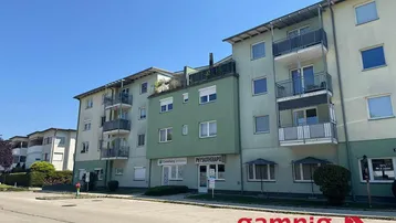 Gebäudeansicht mit mehreren Balkonen und Geschäften im Erdgeschoss unter blauem Himmel.