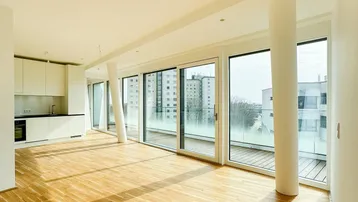 Heller, offener Wohnbereich mit integrierter Küche, Holzboden und großen Fenstern zum Balkon.