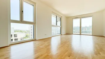 Helles, leeres Zimmer mit großen Fenstern, Holzboden und Zugang zum Balkon.