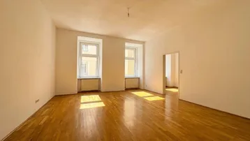 Helles, leeres Zimmer mit Holzboden und zwei großen Fenstern, die viel Sonnenlicht hereinlassen.