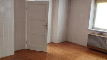 Helles Zimmer mit Holzdielenboden, gemusterter Tapete und einem Fenster mit Heizkörper.