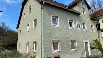 Grünes Gebäude mit traditionellem Ziegeldach und mehreren Fenstern, umgeben von leichter Vegetation.