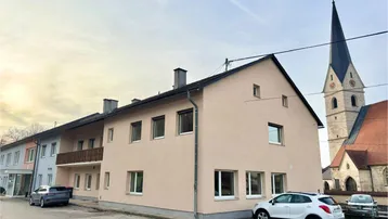 Gebäudeansicht mit beiger Fassade, Satteldach und angrenzender Kirche im Hintergrund.