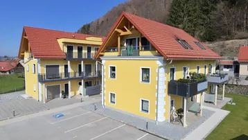 Zwei gelbe Mehrfamilienhäuser mit roten Ziegeldächern, Balkonen und Parkplätzen in ländlicher Umgebung.