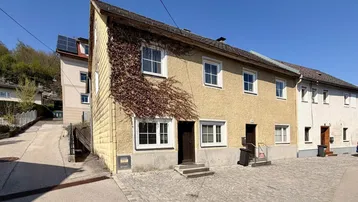 Schräge Ansicht des Hauses mit gelb-beiger Fassade, Dachneigung und gepflastertem Vorplatz.