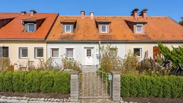 Frontansicht eines Reihenhauses mit Vorgarten und gepflegter Hecke unter blauem Himmel.