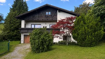Traditionelles Haus mit weißer Fassade, Holzbalkonen und gepflegtem Vorgarten unter blauem Himmel.