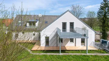 Modernes Haus mit großer Holzterrasse und Balkon, umgeben von grüner Landschaft unter blauem Himmel.