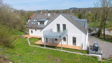 Modernes Haus mit Balkon und Holzterrasse in einer grünen, hügeligen Landschaft.