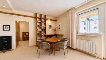 Heller Wohn- und Essbereich mit Teppichboden, integriertem Regal und Blick in ein Schlafzimmer.