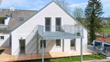 Modernes Haus mit weißer Fassade, großem Balkon und Holzterrasse im Grünen.