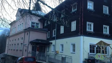 Gebäude mit traditioneller Holzfassade und rosa Anbau, umgeben von Bäumen, mit einer Rampe am Eingang.