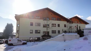 Mehrfamilienhaus im alpinen Stil mit hellbeiger Fassade und Holzbalkonen, umgeben von Schnee und Bergen.