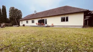 Hausfassade mit großer Rasenfläche und einer Terrasse mit Gartenmöbeln unter bewölktem Himmel.