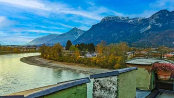 Weitläufiger Blick über einen Fluss und eine Stadt, umgeben von majestätischen Bergen unter blauem Himmel.