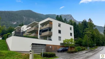 Modernes Apartmentgebäude mit Balkonen und Tiefgarage, umgeben von grünen Bergen.